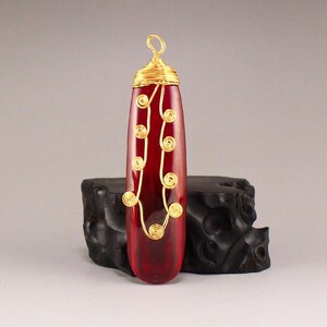 May include: A red gemstone pendant with gold wire wrapped around it. The pendant is shaped like a long oval and has a loop at the top for hanging.