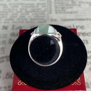 May include: A silver ring with a large, oval-shaped, light green gemstone. The ring is set on a black velvet surface, with a red box visible in the background. The gemstone is the focal point, showcasing its color and shape.