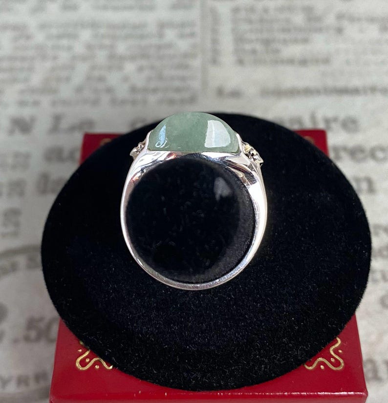 May include: A silver ring with a large, oval-shaped, light green gemstone. The ring is set on a black velvet surface, with a red box visible in the background. The gemstone is the focal point, showcasing its color and shape.