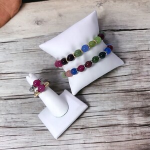 May include: Two colorful beaded bracelets on a white display stand, and a ring with pink and red stones on a white ring stand. The bracelets are made of green, blue, and purple beads.