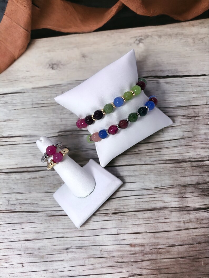 May include: Two colorful beaded bracelets on a white display stand, and a ring with pink and red stones on a white ring stand. The bracelets are made of green, blue, and purple beads.