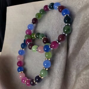 May include: Two colorful beaded bracelets with alternating shades of pink, green, blue, and purple. The beads are separated by silver and gold spacers.