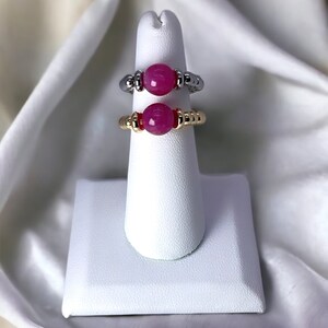 May include: Two rings with pink gemstone beads on a white ring display stand. The ring on the top is silver with gold accents. The ring on the bottom is gold with red accents.