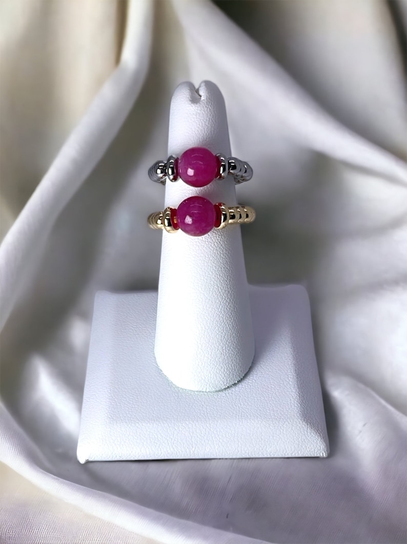May include: Two rings with pink gemstone beads on a white ring display stand. The ring on the top is silver with gold accents. The ring on the bottom is gold with red accents.