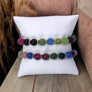 May include: Two bracelets with colorful gemstone beads. The top bracelet has green, blue, and purple beads with gold accents. The bottom bracelet has pink, green, blue, and purple beads with silver accents.