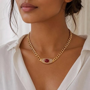 May include: A gold-toned chain link necklace featuring a central oval-shaped pendant. The pendant contains a smooth, deep red cabochon stone surrounded by a border of small, clear, sparkling crystals set into the gold-coloured metal frame.