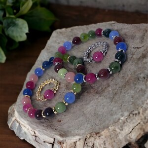 May include: A set of four colorful beaded bracelets with gold accents. The bracelets are made of various shades of blue, green, pink, and purple beads. The bracelets are stacked on top of each other on a wooden surface.