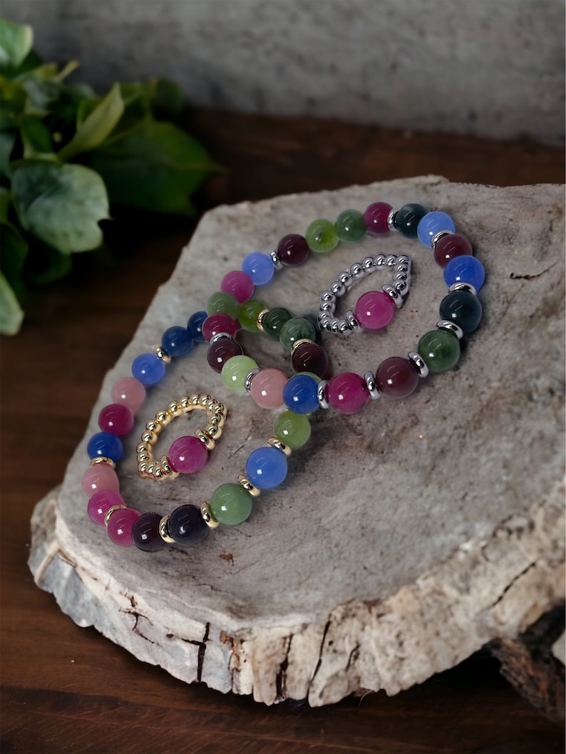 May include: A set of four colorful beaded bracelets with gold accents. The bracelets are made of various shades of blue, green, pink, and purple beads. The bracelets are stacked on top of each other on a wooden surface.
