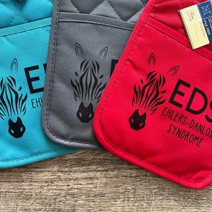 May include: Three pot holders with a zebra print design. The pot holders are teal, gray, and red. The red pot holder has the text "EDS EHLERS-DANLOS SYNDROME" printed on it.