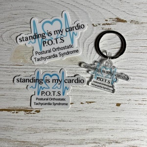 May include: A white keychain with a blue heart and a blue EKG line. The text on the keychain reads "standing is my cardio P.O.T.S. Postural Orthostatic Tachycardia Syndrome".
