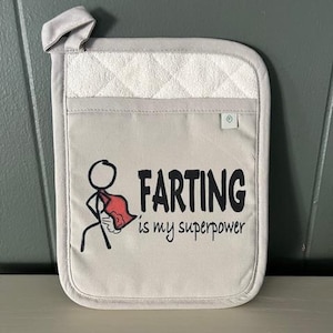 May include: A white cotton pot holder with a black stick figure wearing a red cape and the text "Farting is my superpower".