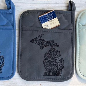 Puede incluir: Tres agarraderas acolchadas con un contorno en blanco y negro del estado de Michigan. Las agarraderas son azules, grises y verde claro.