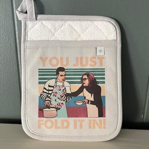 May include: A light gray pot holder with a white quilted top section. The main section features a colorful graphic of two people with the text "YOU JUST FOLD IT IN!" in peach.