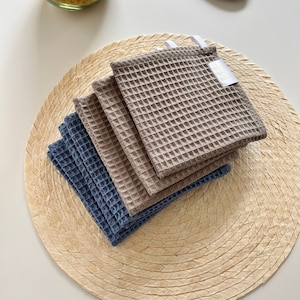 Puede incluir: Un conjunto de cuatro toallas de cocina de tejido de waffle, dos marrones y dos azules, sobre un mantel de paja trenzada. Las toallas tienen una etiqueta blanca con el texto "The Waffle Kitchen" en ellas.