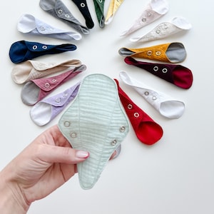 Könnte beinhalten: Eine Auswahl an wiederverwendbaren Stoff-Menstruationseinlagen in verschiedenen Farben und Mustern, darunter Grün, Rot, Gelb, Weiß und Schwarz. Die Einlagen sind in einem Kreis angeordnet, wobei eine Einlage in einer Hand gehalten wird.