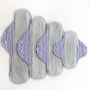 Puede incluir: Cuatro compresas menstruales de tela reutilizables grises con tela a rayas azul y blanca en los bordes. Las compresas están dispuestas en orden descendente de tamaño.