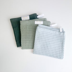 May include: Three waffle-weave washcloths in shades of green and blue. The washcloths have a square shape with a textured surface. Each cloth has a white tag with gold text. The washcloths are arranged on a white surface.