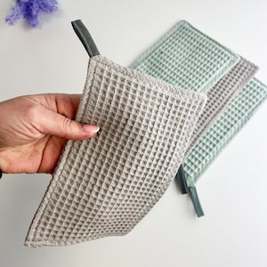 May include: Three waffle weave kitchen towels with hanging loops. One towel is beige, and two are light green.