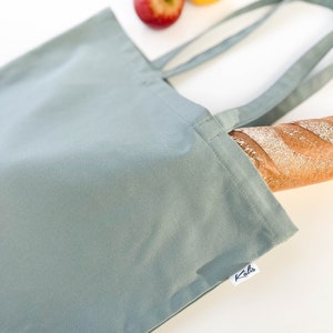 May include: A sage green canvas tote bag with a white label that says "Kaha" on it. The bag is lying on a white surface with a loaf of bread and apples in the background.