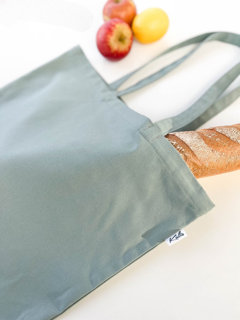 May include: A sage green canvas tote bag with a white label that says "Kaha" on it. The bag is lying on a white surface with a loaf of bread and apples in the background.