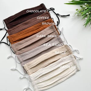 Può includere: Una collezione di mascherine plissettate in vari colori neutri, tra cui cioccolato, caffè, marrone, beige scuro, sabbia marina, beige e vaniglia. Ogni mascherina ha elastici regolabili ed è etichettata con il nome del colore.