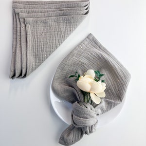 Peut inclure: Une serviette en lin gris avec des paillettes dor&eacute;es, pli&eacute;e et nou&eacute;e avec une fleur blanche, sur une assiette blanche.
