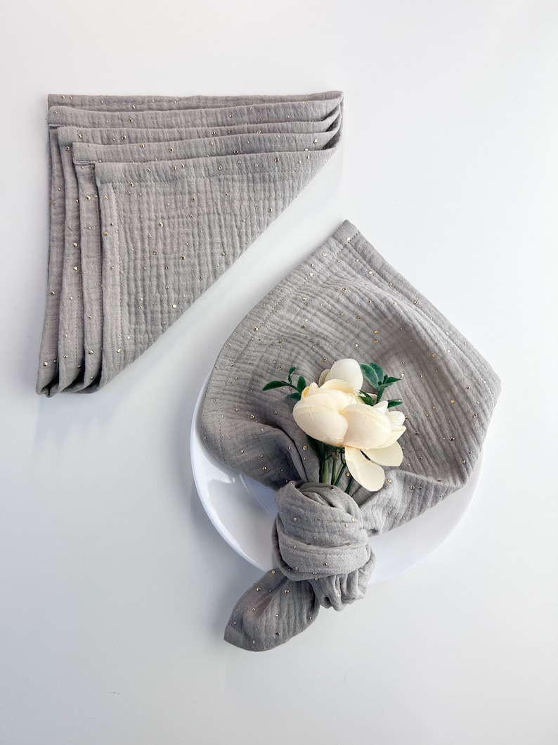 Peut inclure: Une serviette en lin gris avec des paillettes dor&eacute;es, pli&eacute;e et nou&eacute;e avec une fleur blanche, sur une assiette blanche.