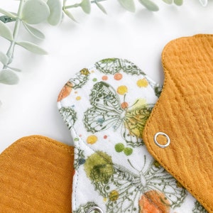 May include: Two reusable cloth menstrual pads. One pad is white with a butterfly print and the other is a solid mustard yellow. Both pads have a snap closure.