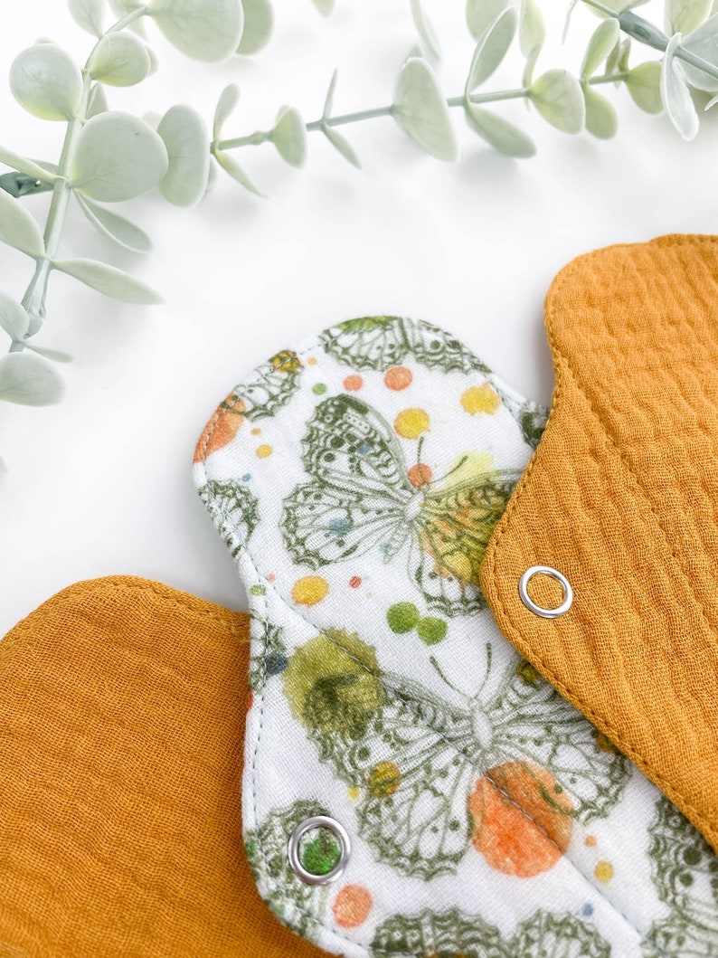 May include: Two reusable cloth menstrual pads. One pad is white with a butterfly print and the other is a solid mustard yellow. Both pads have a snap closure.