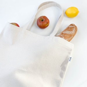 May include: A white canvas tote bag with a brown baguette, a yellow lemon, and two red apples inside. The bag has a black label with the word "Kola" on it.