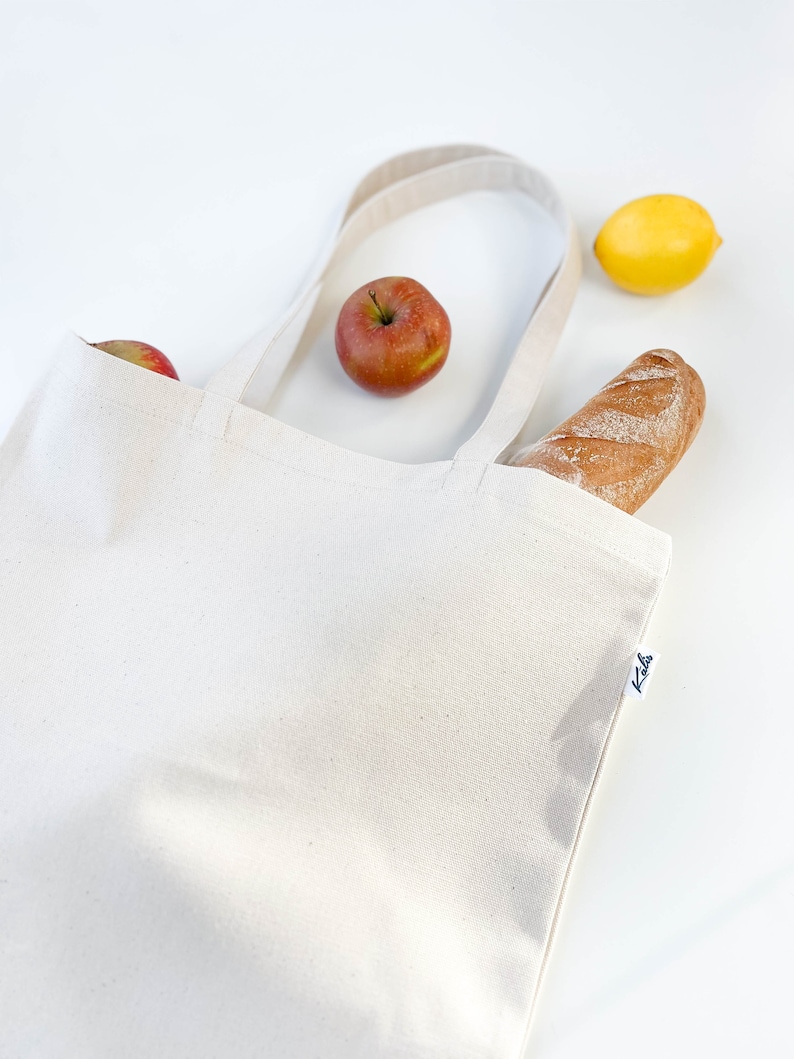 May include: A white canvas tote bag with a brown baguette, a yellow lemon, and two red apples inside. The bag has a black label with the word "Kola" on it.