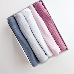Peut inclure: Cinq linges en mousseline roul&eacute;s dans une bo&icirc;te blanche. Les linges sont dans des tons de bleu, gris, blanc, rose et rose.