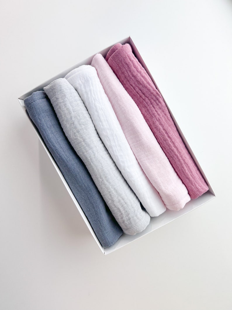 Peut inclure: Cinq linges en mousseline roul&eacute;s dans une bo&icirc;te blanche. Les linges sont dans des tons de bleu, gris, blanc, rose et rose.