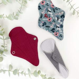 May include: Three reusable cloth menstrual pads in different colours and patterns. A red pad, a grey pad, and a blue pad with a floral pattern.