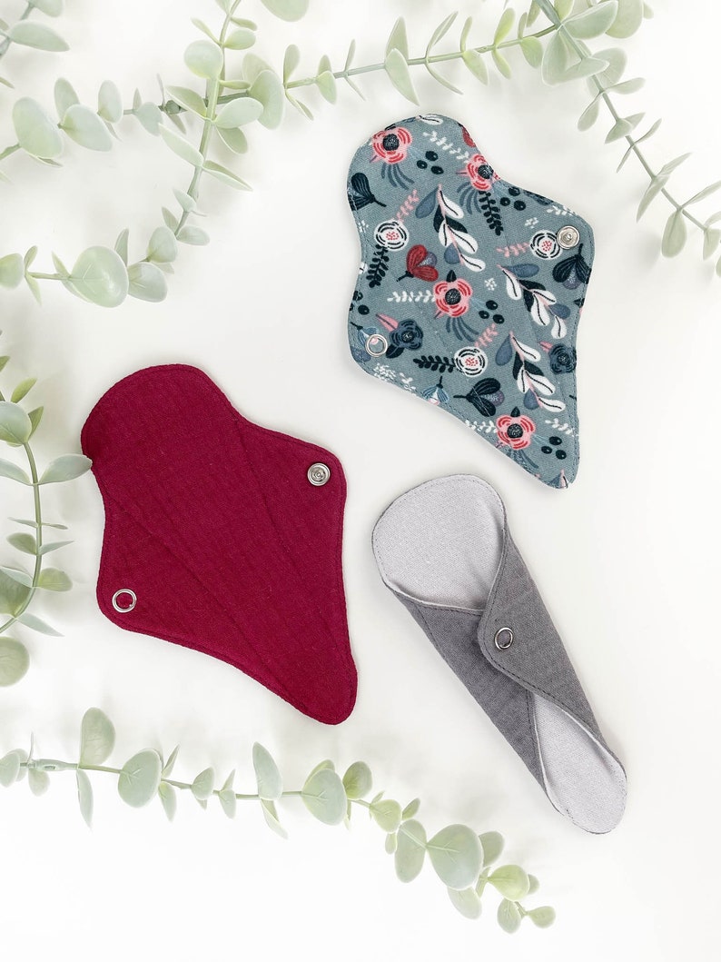 May include: Three reusable cloth menstrual pads in different colours and patterns. A red pad, a grey pad, and a blue pad with a floral pattern.