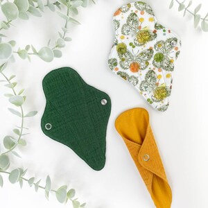 May include: Three reusable cloth menstrual pads in different colours and patterns. A green pad, a yellow pad, and a white pad with a butterfly print.