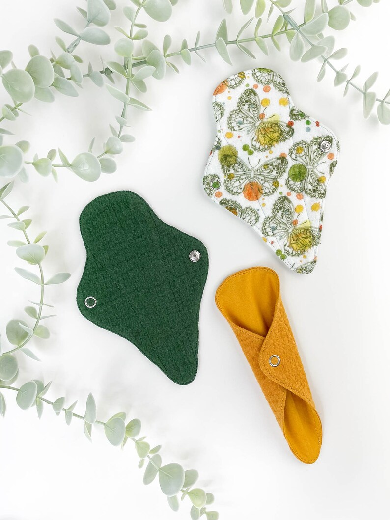 May include: Three reusable cloth menstrual pads in different colours and patterns. A green pad, a yellow pad, and a white pad with a butterfly print.