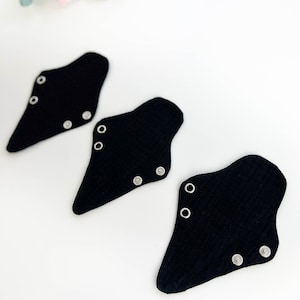 Peut inclure: Trois serviettes hygiéniques réutilisables noires en tissu avec des fermetures à pression. Les serviettes sont de forme triangulaire avec des bords arrondis. Le fond est blanc avec une branche de fleurs roses.