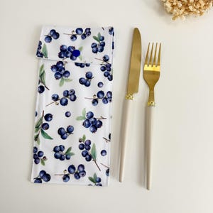May include: A white cutlery pouch featuring a blueberry design, alongside a gold knife and fork with cream handles. The pouch is secured with a blue button. The blueberry pattern includes blue berries and green leaves.