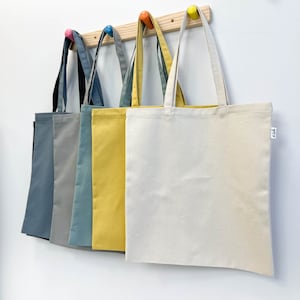 Puede incluir: Seis bolsas de mano de lona en varios colores, incluyendo azul, gris y amarillo, colgadas de un estante de madera con clavijas de colores. Las bolsas tienen asas largas y un diseño sencillo y minimalista, adecuadas para llevar la compra o artículos personales.