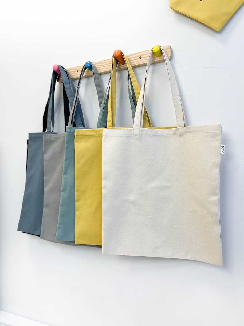 May include: Five canvas tote bags hanging on a wooden rack. The bags are in various colors, including black, gray, teal, yellow, and natural. The bags have a simple design and are perfect for carrying everyday essentials.