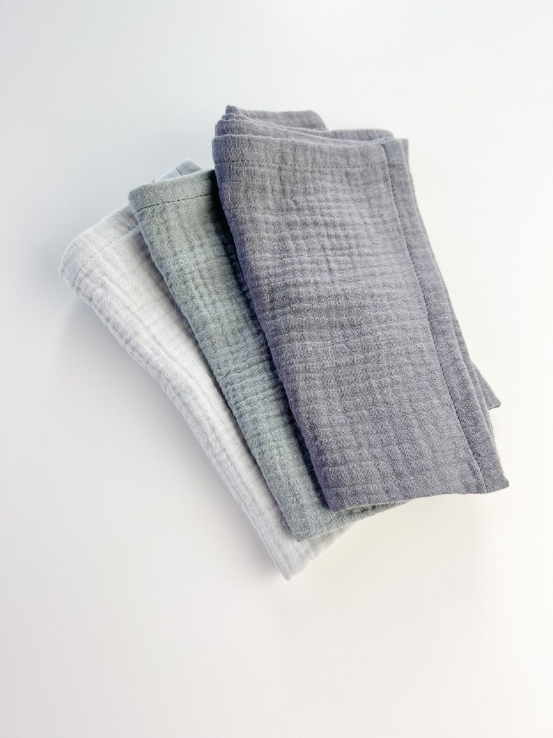 Peut inclure: Trois linges en mousseline de coton pli&eacute;s ensemble. Le linge du dessus est gris fonc&eacute;, le linge du milieu est gris clair et le linge du dessous est blanc.