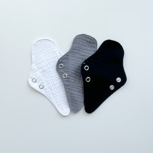 May include: Three baby bibs in white, gray, and black, each with snap closures. The bibs are made of a soft, textured fabric and are arranged on a white surface. The bibs are designed to catch spills and drool.