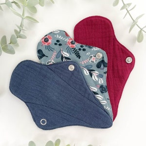 May include: Three reusable cloth menstrual pads in different colours and patterns. The pads are folded and arranged in a fan shape. The top pad is a solid red colour, the middle pad has a floral pattern, and the bottom pad is a solid dark blue colour.