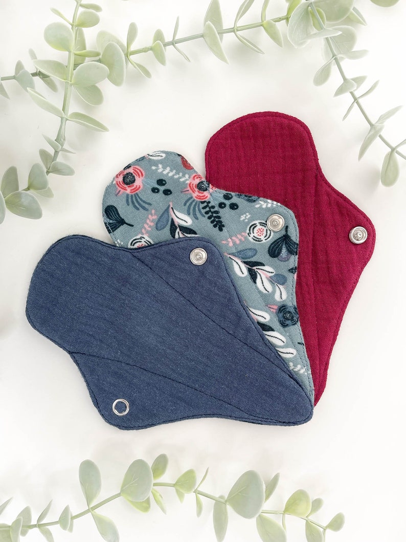 May include: Three reusable cloth menstrual pads in different colours and patterns. The pads are folded and arranged in a fan shape. The top pad is a solid red colour, the middle pad has a floral pattern, and the bottom pad is a solid dark blue colour.