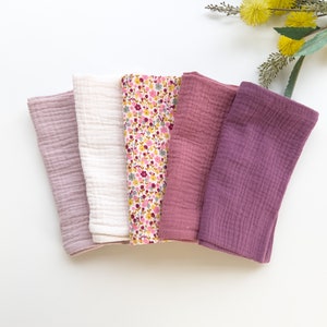 May include: Five soft, cotton muslin cloths in shades of pink and purple. The cloths are folded and arranged in a row on a white surface.