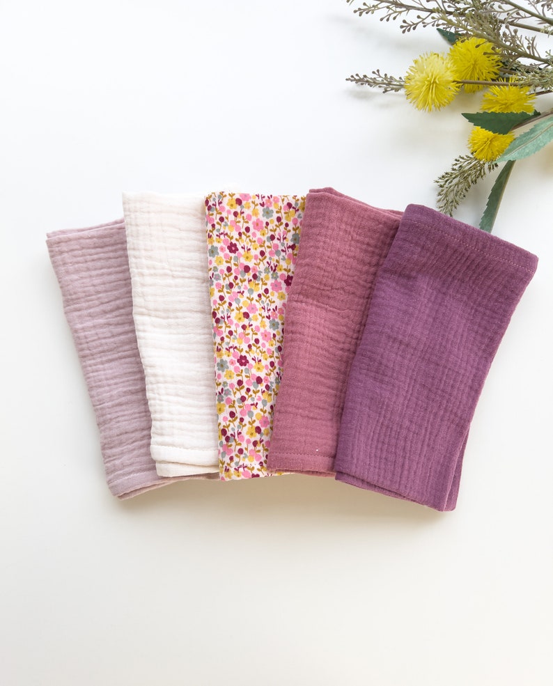 Peut inclure: Cinq linges en mousseline de coton doux dans des tons de rose et de violet. Les linges sont pli&eacute;s et dispos&eacute;s en rang&eacute;e sur une surface blanche.