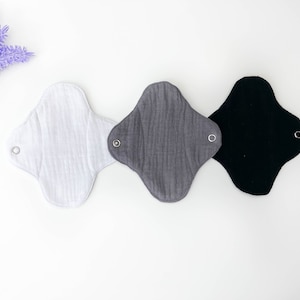 May include: Three reusable cloth menstrual pads in white, gray, and black. Each pad has a snap closure on each side.