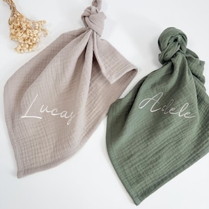 Può includere: Due bavaglini annodati in mussola per neonati, uno color talpa e uno verde oliva, ciascuno ricamato con un nome in corsivo. Il bavaglino color talpa legge "Lucas" e quello verde legge "Adele".