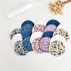 Può includere: Una collezione di dischetti per il viso riutilizzabili in vari modelli e colori. I dischetti includono denim, motivi floreali e spugna bianca. Una bottiglia bianca, un contagocce e fiori secchi sono anche nell'immagine.